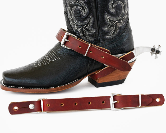 A pair of brown fold-back spur straps on a black boot, with silver buckle and hardware.