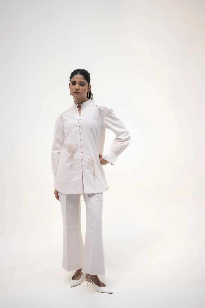 A model standing with a hand on her hip, wearing a long white shirt and white trousers, in a neutral pose against a soft background.