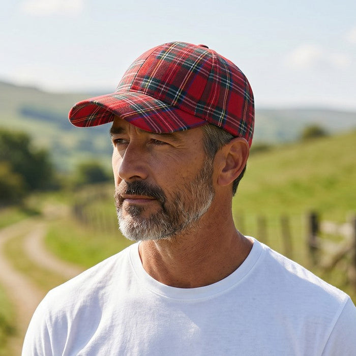 A man wearing a red and black plaid baseball cap and a plain white t-shirt, standing outdoors on a dirt path with green hills in the background.